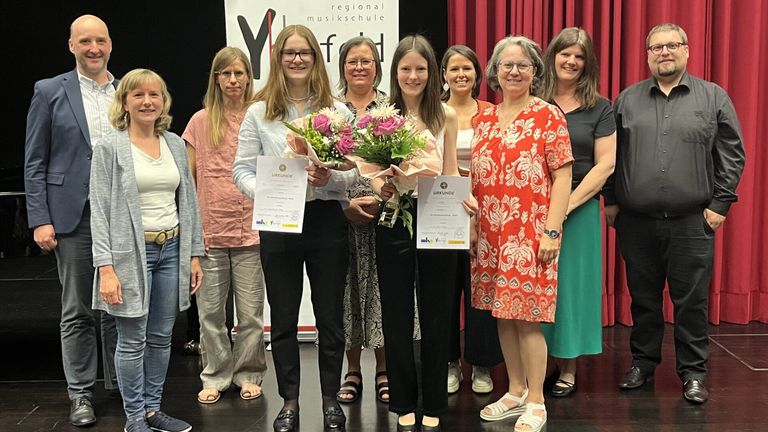 Eine Gruppe von Frauen steht zusammen, lächelt und hält Urkunden. Sie sind alle lässig gekleidet. Eine Frau in der Mitte hält einen Blumenstrauß. Hinter ihnen hängt ein roter Vorhang.