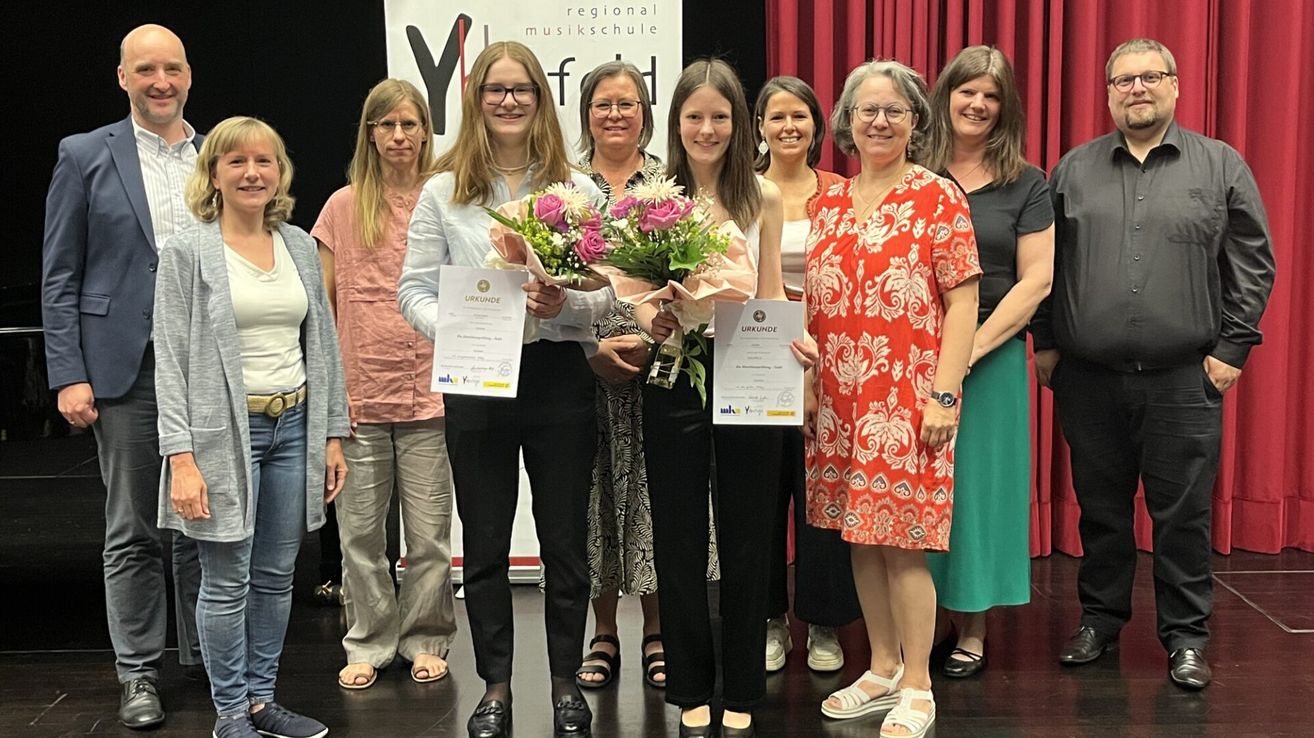 Eine Gruppe von Frauen steht zusammen, lächelt und hält Urkunden. Sie sind alle lässig gekleidet. Eine Frau in der Mitte hält einen Blumenstrauß. Hinter ihnen hängt ein roter Vorhang.