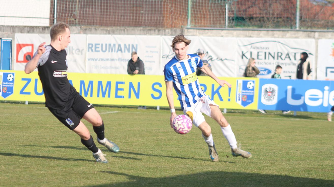 Fußballspieler im blau-weißen Trikot tritt einen rosa Ball auf einem Feld, ein weiterer Spieler im schwarzen Trikot im Vordergrund.