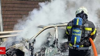 Ein Feuerwehrmann mit Helm und blauem Rucksack mit der Aufschrift 'Dräger' steht neben einem ausgebrannten Auto, aus dem Rauch aufsteigt.