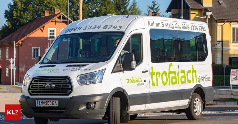 Ein weißer Van mit der Aufschrift 'trofairach' fährt auf einer Straße mit einem Mann im Inneren, Kennzeichen '955 BA'. Bäume und Gebäude sind im Hintergrund sichtbar.
