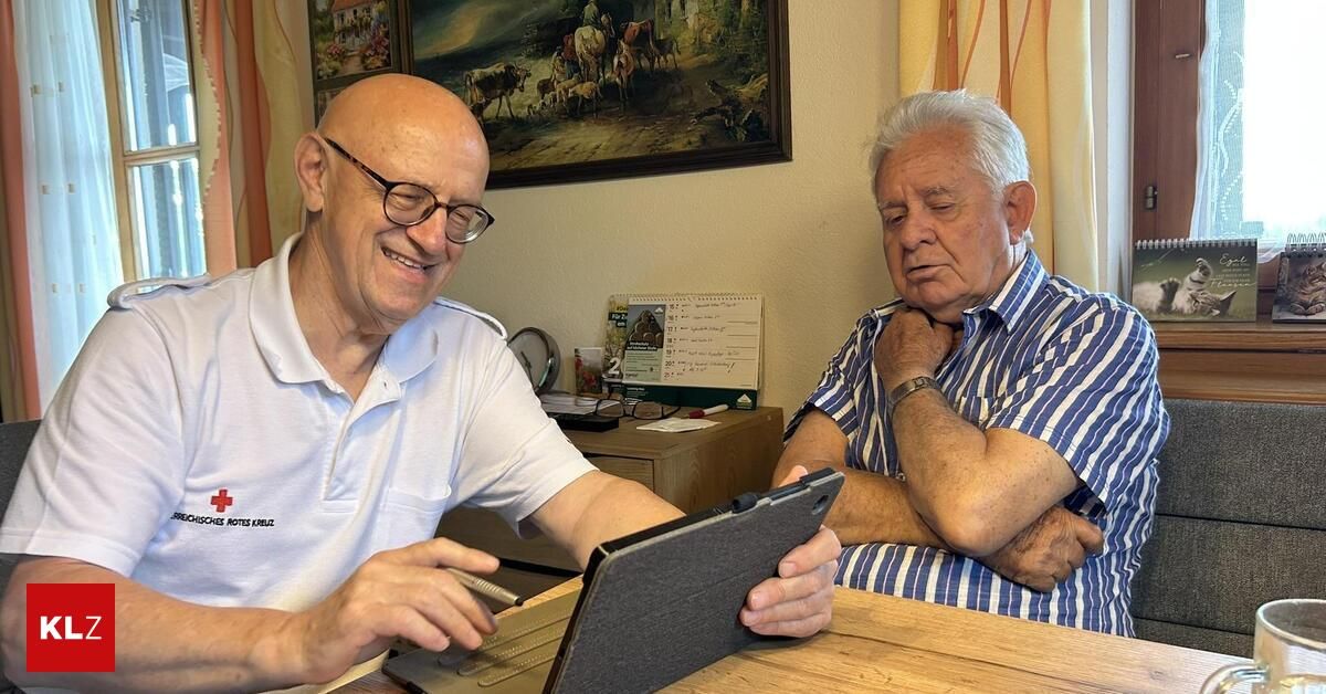 Zwei ältere Männer sitzen an einem Tisch, einer lächelt und benutzt ein Tablet, während der andere die Arme verschränkt hat und wegschaut. Hinter ihnen ist eine Wand mit einem Gemälde von Tieren.