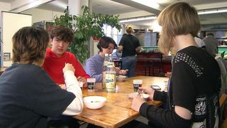 Eine Gruppe junger Leute sitzt um einen Holztisch und isst. Ein Mädchen trägt eine schwarze Lederjacke. Eine Pflanze steht hinter ihnen, und eine Frau in einem schwarzen Shirt geht im Hintergrund.