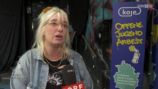 Eine Frau mit blonden Haaren, Brille und Jeansjacke spricht in ein Mikrofon vor einer Graffiti-Wand. Ein Schild steht für 'ORF'.