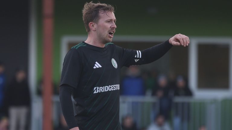 Ein Mann in einem schwarzen Fußballtrikot schreit und zeigt nach rechts während eines Spiels. Er trägt einen grün-schwarz gestreiften Kragen.