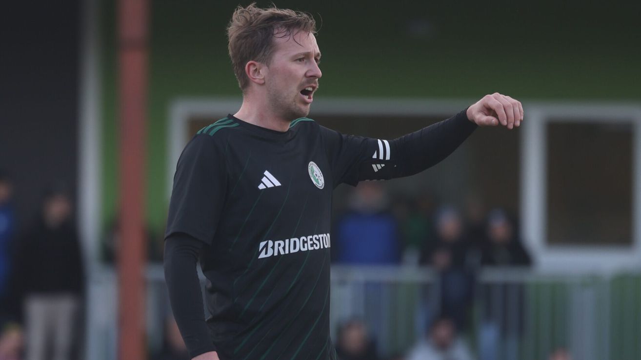 Ein Mann in einem schwarzen Fußballtrikot schreit und zeigt nach rechts während eines Spiels. Er trägt einen grün-schwarz gestreiften Kragen.