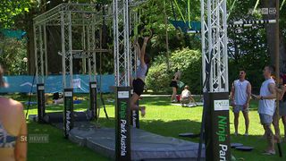 Eine Frau führt einen Klimmzug an einer Stange in einem Park durch, andere Personen sind in der Umgebung, einige sitzen auf dem Rasen.