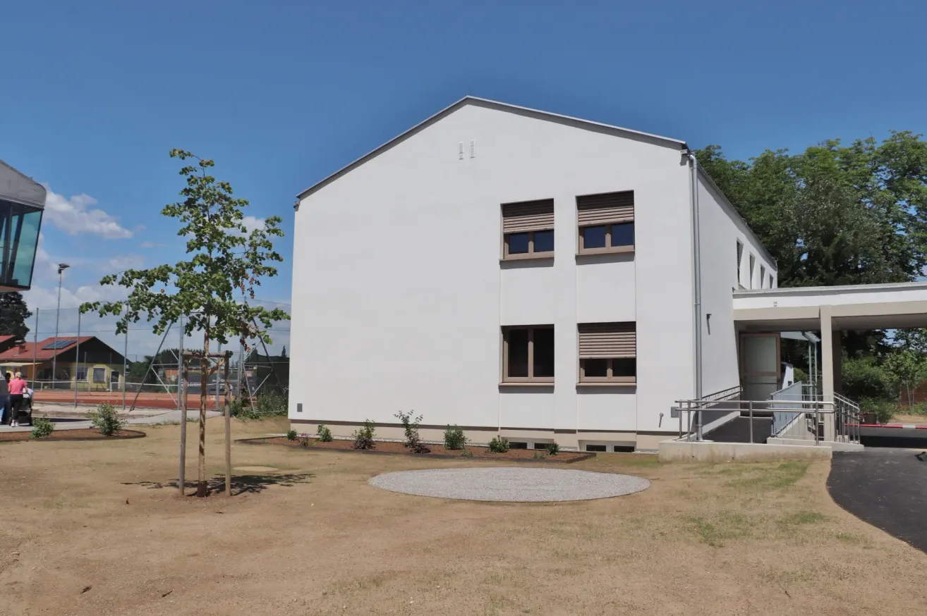 Ein weißes Gebäude mit mehreren Fenstern und einem Baum steht in einem offenen Bereich. Das Gebäude hat eine kleine runde Betonplattform vor sich.