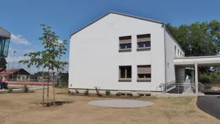 Ein weißes Gebäude mit mehreren Fenstern und einem Baum steht in einem offenen Bereich. Das Gebäude hat eine kleine runde Betonplattform vor sich.