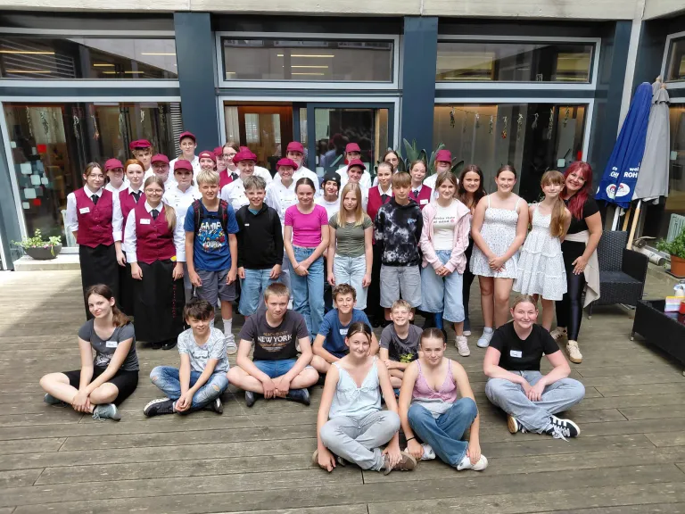 Eine Gruppe von Menschen, meist junge Erwachsene, posiert für ein Foto auf einer Holzterrasse vor einem Gebäude. Sie tragen Freizeitkleidung, einige mit Hüten.