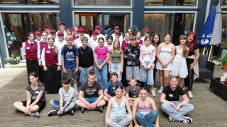 Eine Gruppe von Menschen, meist junge Erwachsene, posiert für ein Foto auf einer Holzterrasse vor einem Gebäude. Sie tragen Freizeitkleidung, einige mit Hüten.