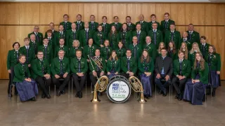 Eine Gruppe von Musikern in grünen Uniformen und Krawatten posiert für ein Foto mit Instrumenten vor einer Holzwand. Ein Schlagzeug mit dem Text 'MUSIK FEST 2026' steht in der Mitte.