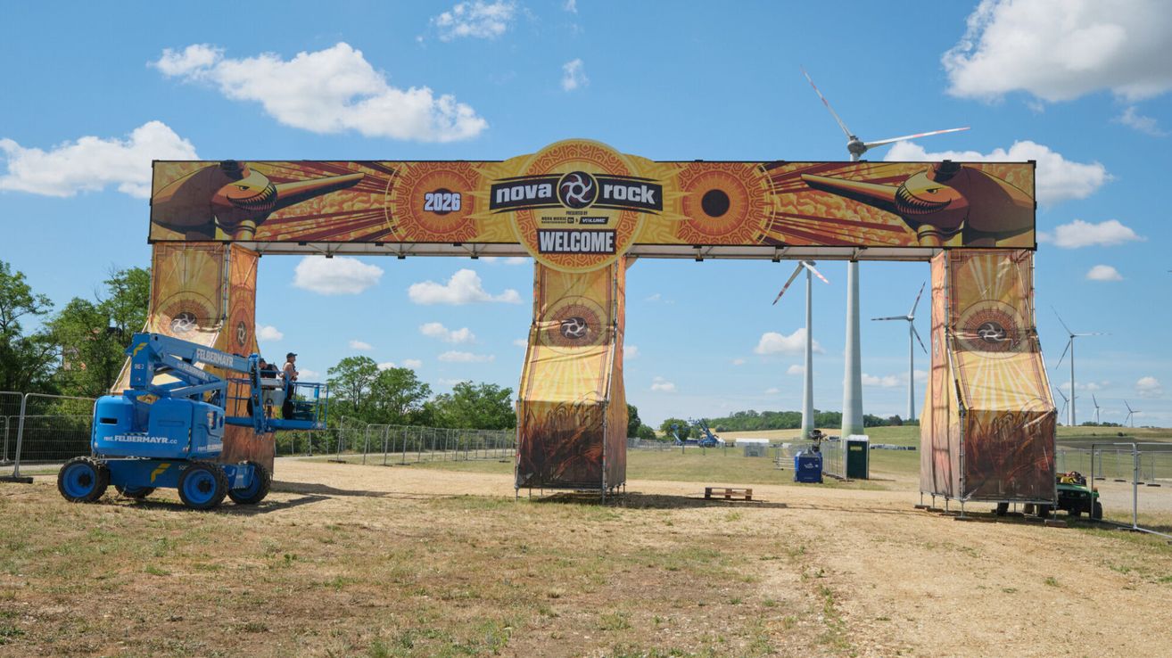 Ein farbenfroher Willkommensbogen für das Nova Rock Festival 2026. Er steht auf einem Feld, mit Windrädern in der Ferne unter einem blauen Himmel.