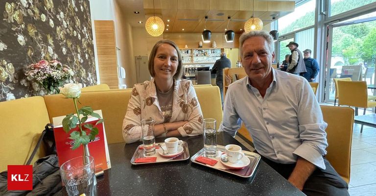 Ein Mann und eine Frau sitzen an einem Tisch in einem Restaurant mit Kaffeetassen und Untertassen. Die Frau lächelt und hat ein Armband am Handgelenk.