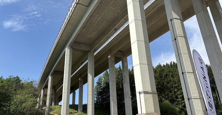 Eine Betonbrücke mit mehreren Pfeilern, die sich über einem Grasbereich erhebt, mit Bäumen im Hintergrund unter einem blauen Himmel.