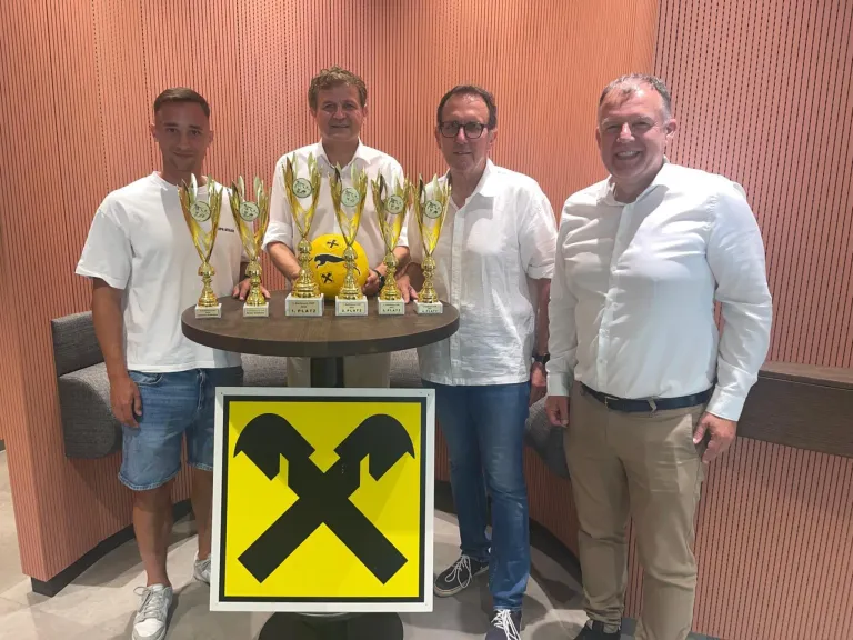 Fünf Männer stehen hinter einem Tisch mit mehreren Trophäen. Der Mann auf der linken Seite trägt Shorts. Der Tisch hält einen gelben Fußball. Ein gelbes und schwarzes Schild ist im Vordergrund.