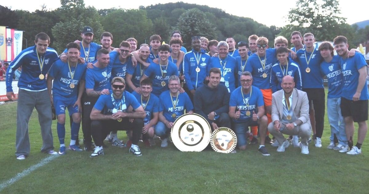 Eine Gruppe von Männern in blauen Trikots posiert für ein Foto mit Medaillen und einer Trophäe auf einem Rasenfeld.