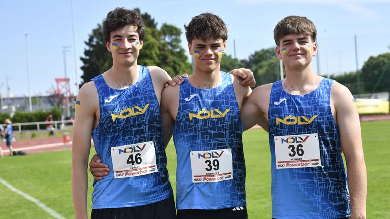 Three young men in blue NOLV jerseys stand together in a field. They have numbers 46, 39, and 36 on their jerseys. They are smiling and have yellow paint on their faces.