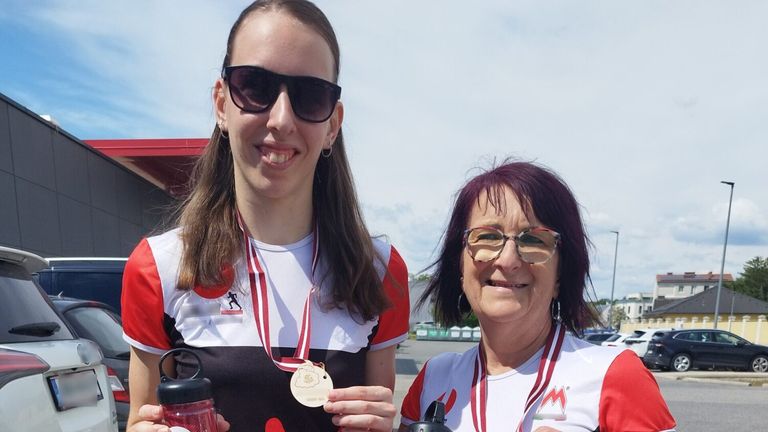Eine Frau mit einer Medaille um den Hals lächelt für ein Foto mit einer anderen Frau auf einem Parkplatz. Sie tragen beide eine Sonnenbrille und halten Wasserflaschen.