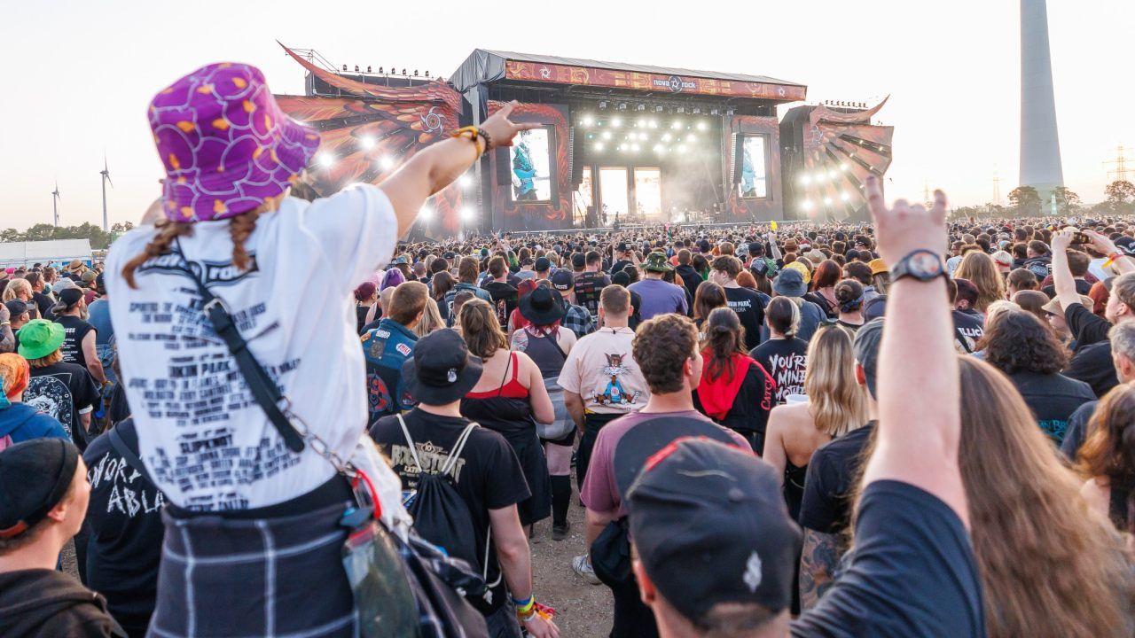 Eine Menschenmenge versammelt sich für ein Musikfestival mit einer großen Bühne, mehreren Scheinwerfern und Künstlern. Einige Leute tragen Hüte, Rucksäcke und Armbänder.