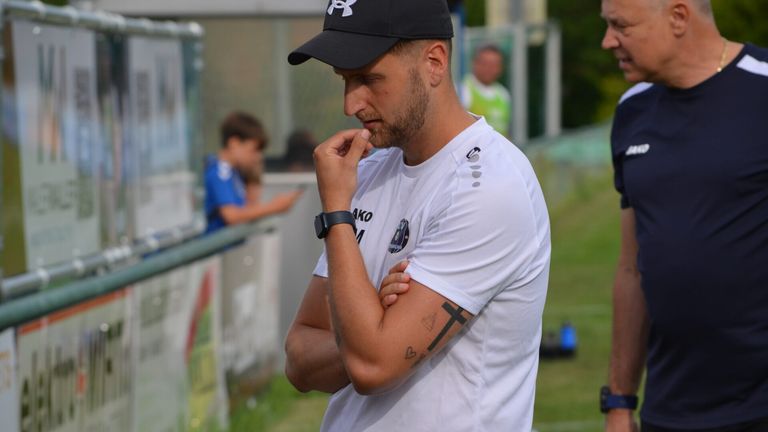 A man wearing a black hat and a white Under Armour shirt watches a soccer game, holding his chin with his right hand.
