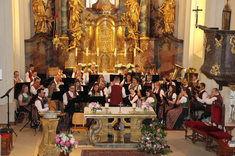 Ein Blasorchester spielt in einer Kirche. Die Bandmitglieder sitzen und spielen ihre Instrumente, während Kerzen und Blumen den Altar schmücken.