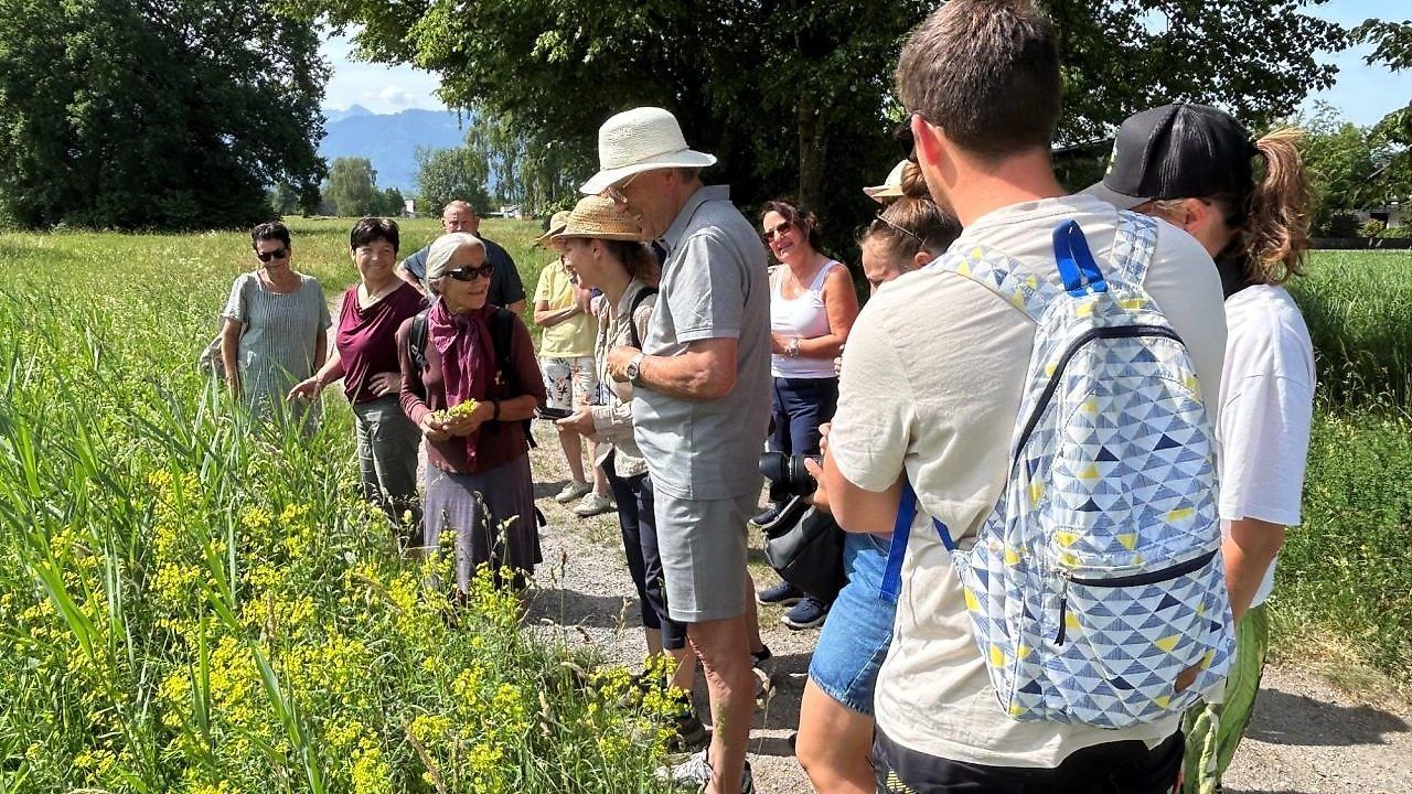 Eine Gruppe von Leuten in Hüten steht in einem Feld mit hohem Gras. Einige schauen einen Mann mit einer Kamera an.