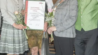 Vier Personen stehen in einer Gruppe, zwei halten Blumensträuße, ein Mann hält ein Zertifikat und sie scheinen alle eine Leistung zu feiern.