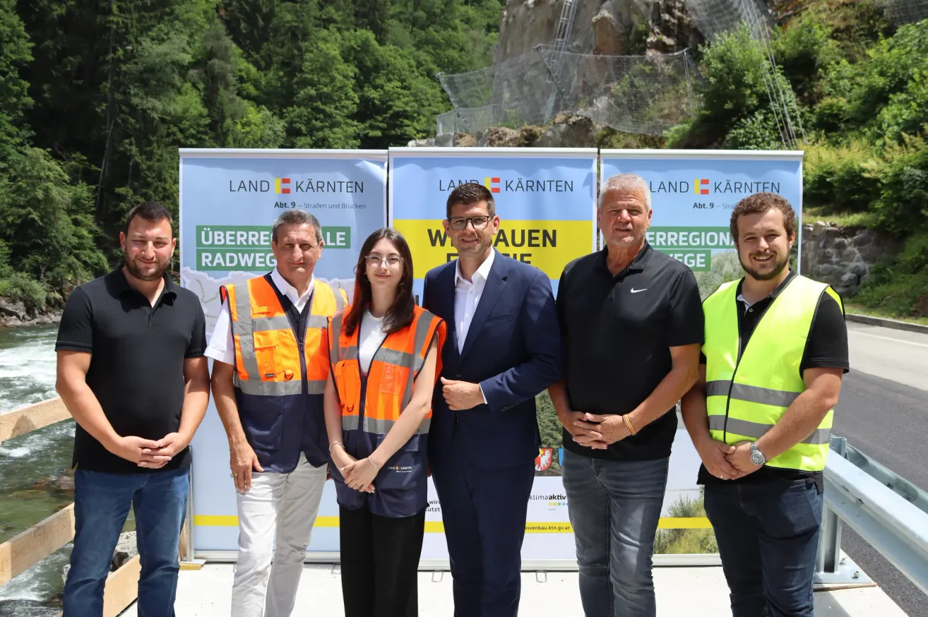 Fünf Personen stehen vor Plakaten mit dem Text 'Land Kärnten' und posieren für ein Foto. Der Mann in der Mitte trägt eine Brille und einen Anzug. Rechts neben ihm steht ein Mann in einem schwarzen T-Shirt und Jeans, links ein Mann in einer gelben Weste. Dahinter befindet sich eine Steinmauer und Bäume.