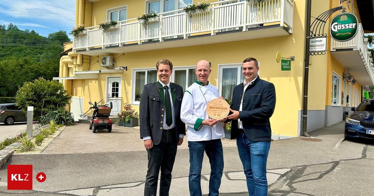 Drei Männer stehen vor einem gelben Gebäude, zwei halten eine Plakette mit deutschem Text. Der Mann links trägt ein grünes Band. Der Mann in der Mitte trägt ein Kochoutfit. Der Mann rechts trägt einen Anzug. Ein Roller steht links geparkt.