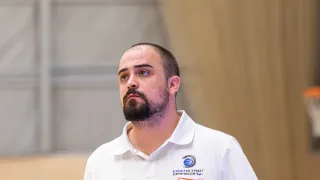Ein Mann in einem weißen Hemd mit Logo steht und schaut zur Seite, möglicherweise in einer Sportarena.