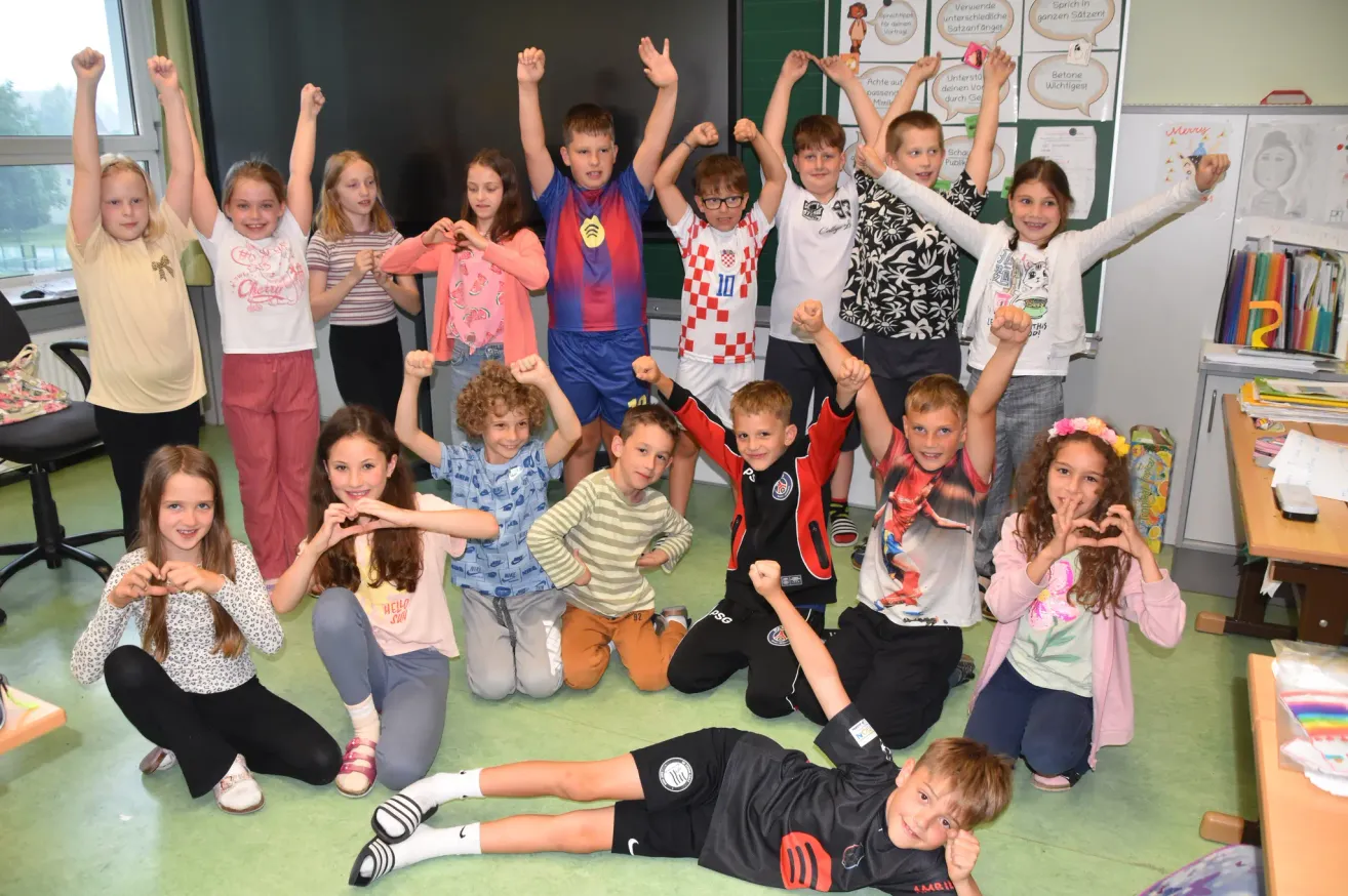 A group of children and young adults are posing for a photograph inside a classroom. They are smiling and have their hands raised. They are dressed in various colored shirts and pants. Some are kneeling, while others are sitting on the floor.