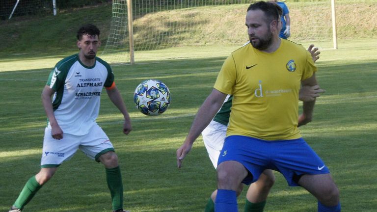 Two soccer players are in the middle of a match. One player is wearing a green and white uniform and the other is in a yellow and blue uniform. A soccer ball is in the air.