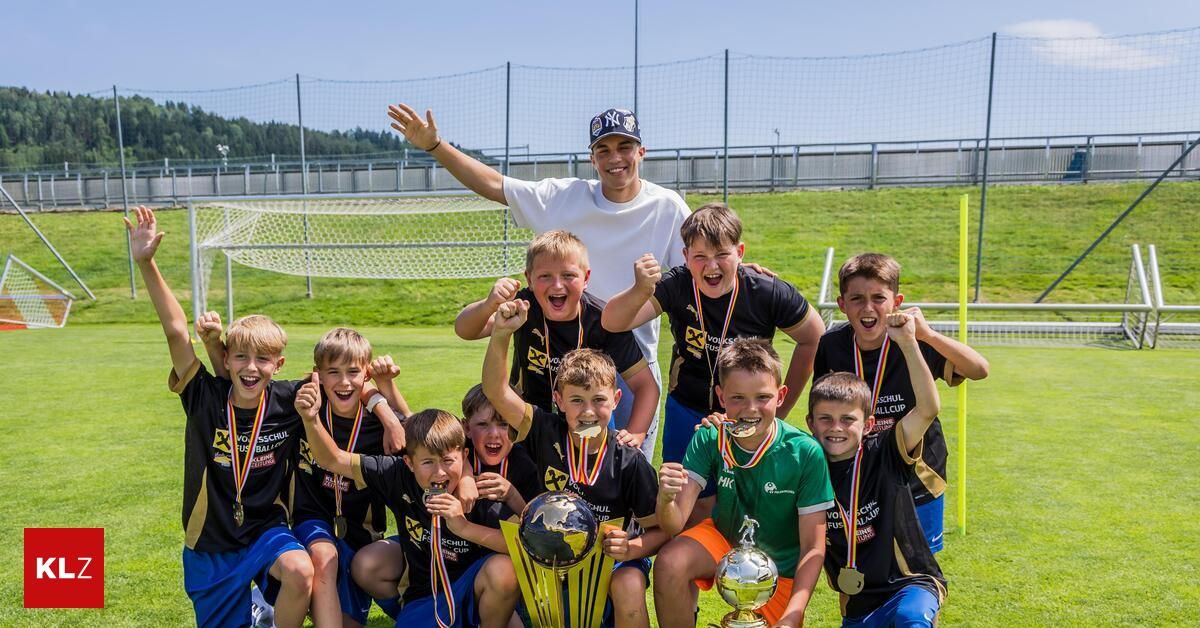 Eine Gruppe junger Jungen in Sportuniformen steht mit ihrem Trainer auf einem Feld und hält Trophäen. Sie alle lächeln und einige heben die Hände. Der Trainer trägt einen Hut und ein weißes T-Shirt. Im Hintergrund befindet sich ein Tornetz.