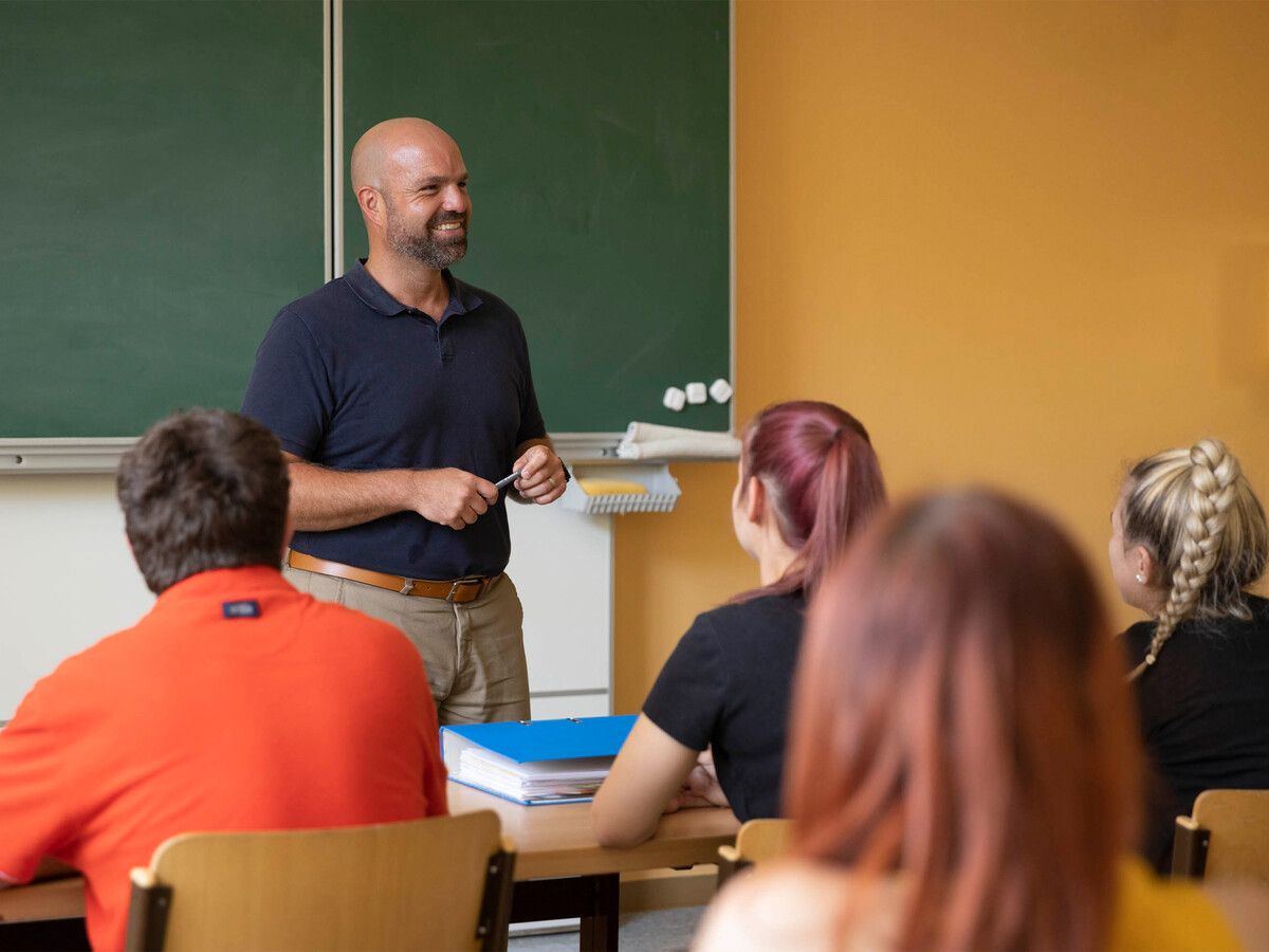 Ein männlicher Lehrer steht vor einem Klassenzimmer, lächelt und hält einen Stift. Er ist von Schülern umgeben, von denen einige ihn ansehen.