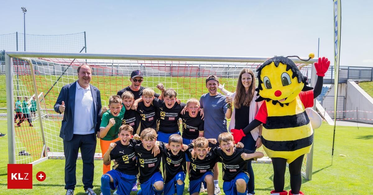 Eine Gruppe von Kindern in passenden Trikots posiert für ein Foto vor einem Fußballtor. Ein Maskottchen in einer Bienenkostüm gesellt sich zu ihnen, mit lächelnden Erwachsenen im Hintergrund.
