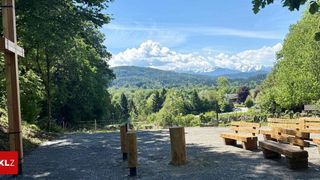 Ein Freiluftbereich mit Holzbänken unter einem sonnigen Himmel. Bäume und Berge sind im Hintergrund.