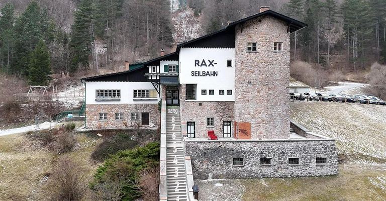An aerial view of the Rax-Seilbahn building with stone walls, multiple windows, and a staircase. The building is set against a backdrop of trees.