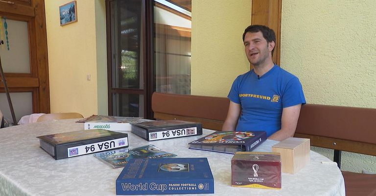 A man in a blue shirt sits at a table with various soccer memorabilia, including Panini World Cup collections.