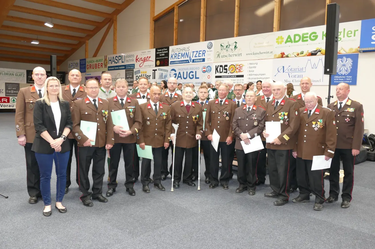 Eine Gruppe von Männern in Militäruniformen posiert für ein Foto. Sie stehen in einer Reihe und halten Papiere. An der Wand hinter ihnen hängen verschiedene Poster und Logos.
