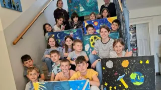 Eine Gruppe von Kindern und Erwachsenen posiert auf einer Treppe für ein Foto und hält bunte Gemälde mit Weltraumthemen hoch.