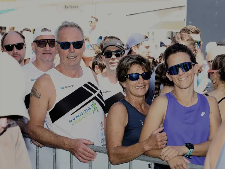 A group of runners are posing for a photo, wearing sunglasses and athletic clothing. A man with a tattoo on his arm stands behind a fence.