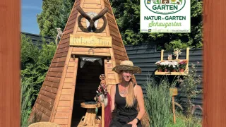Eine Frau sitzt in einer Holz-Tipi mit einem Schild, das Ritual Tipi sagt. Dahinter ist ein Garten mit Pflanzen und einer Holzkiste mit Blumen. Sie hält einen Stock und eine kleine Figur.
