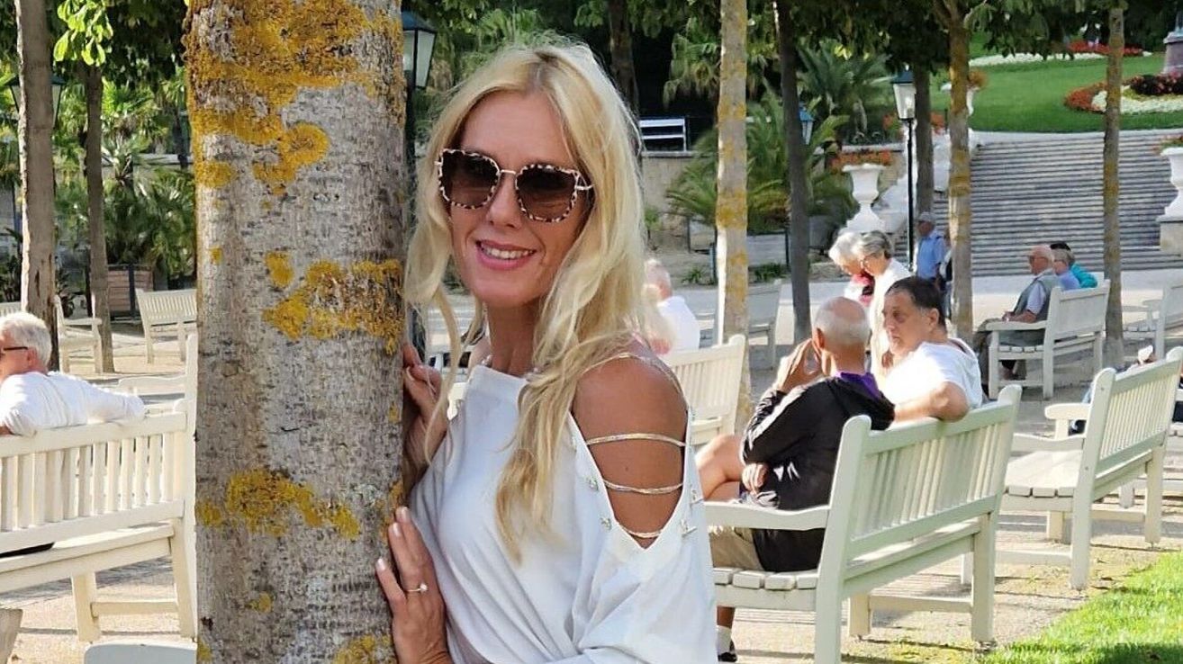 Eine Frau mit blonden Haaren posiert für ein Foto in einem Park in der Nähe eines Baumes, während andere Menschen auf Bänken sitzen.