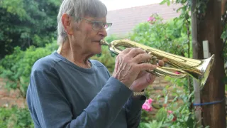 Eine ältere Frau mit Brille und einem blauen Langarmshirt spielt eine goldene Trompete in einem Garten. Hinter ihr sind verschiedene Pflanzen und Bäume.