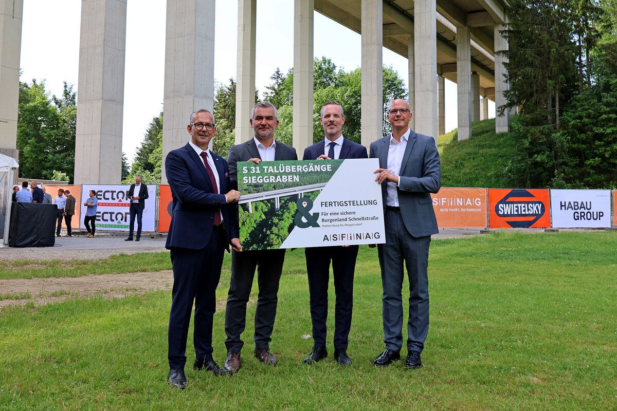 Vier Männer in Anzügen halten ein Schild zur Fertigstellung des S31 Tullberg-Tunnels. Im Hintergrund befindet sich eine Betonbrücke und grüne Bäume.