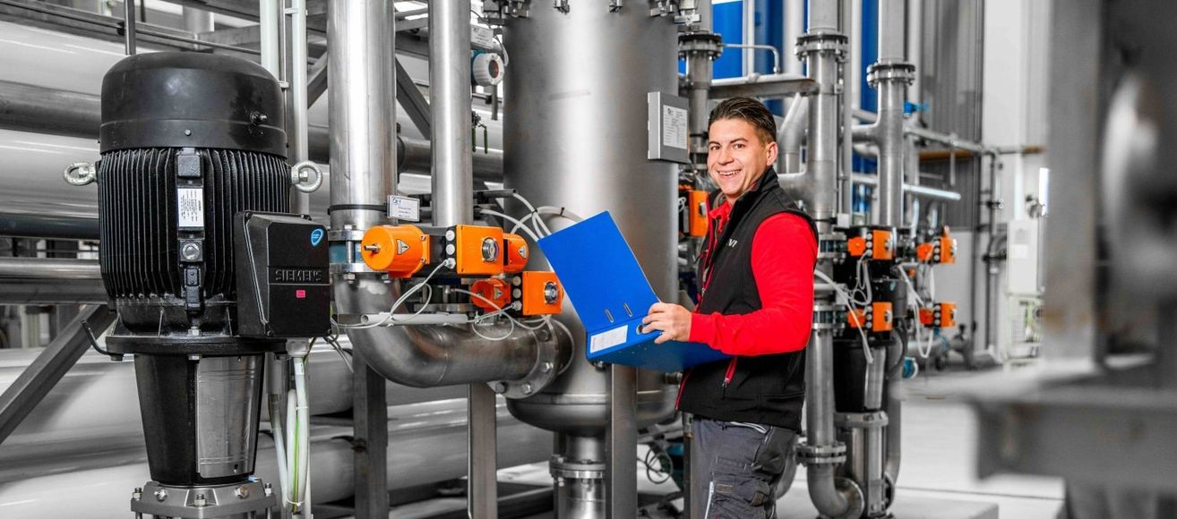 Ein Mann steht in einer industriellen Anlage, lächelt und hält eine blaue Mappe in der Hand, mit Metallrohren und Maschinen im Hintergrund.