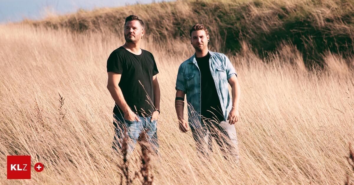 Zwei Männer stehen in einem hohen Grasfeld unter klarem Himmel, einer trägt ein schwarzes T-Shirt und der andere eine Jeansjacke.