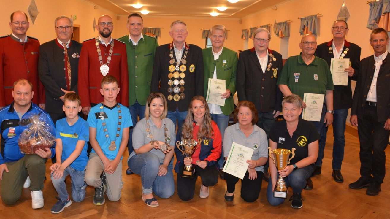 Eine Gruppe von Menschen, möglicherweise Gewinner, posiert für ein Foto, während sie Trophäen und Urkunden in der Hand halten. Einige von ihnen tragen Medaillen und grüne Jacken. Sie befinden sich in einem Raum mit Holzboden und Lampen an der Decke.