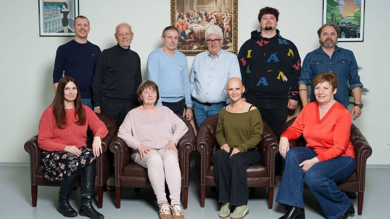 Eine Gruppe von Menschen sitzt auf braunen Stühlen, lächelt und posiert für ein Foto mit einem großen Gemälde im Hintergrund.